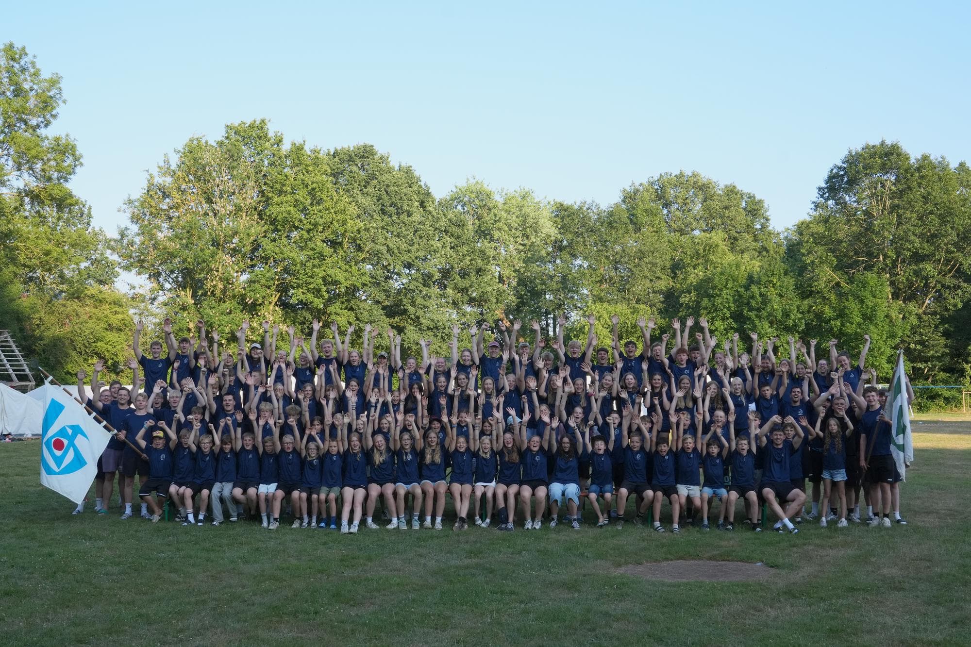 Gruppenfoto vom Zeltlager 2026: alle Kinder und Gruppenleitungen jubeln mit erhobenen Armen auf der Wiese, links und rechts wehen Fahnen